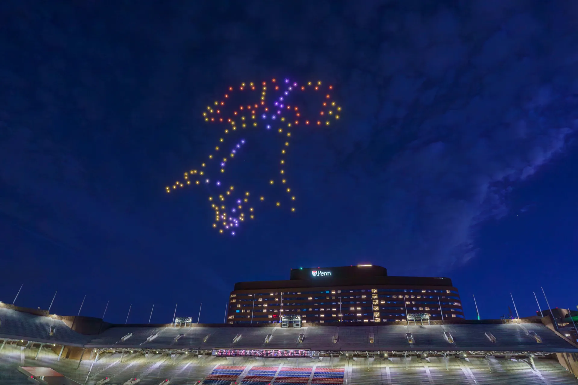 A multicolored drone light formation above Franklin Field with a Penn campus building behind