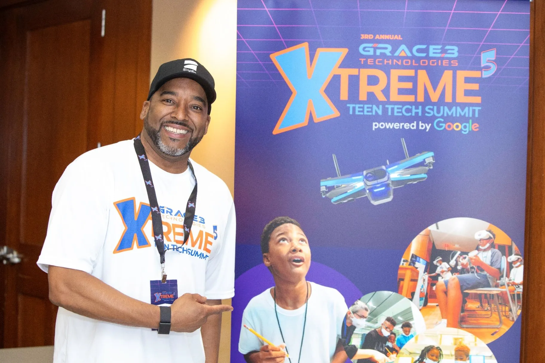 A smiling mentor in a white Xtreme Teen Tech Summit shirt and lanyard pointing beside an Xtreme Teen Tech Summit banner